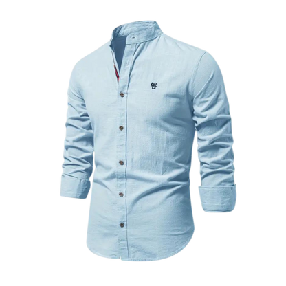 Arkadya Outlet Koszula męska kołnierz stójka długi rękaw krój regularny to jasnoniebieska koszula z zapięciem na guziki z przodu i haftowanym logo, idealna do codziennego użytku, pokazana na rozmytym tle wnętrza.