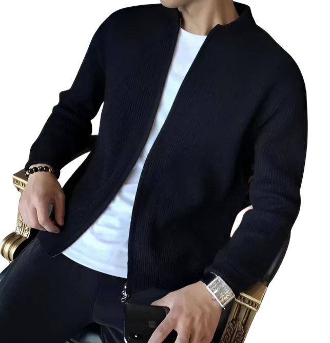 Osoba siedząca na krześle ma na sobie Arkadya Outlet Męski kardigan | Zapinany Na Zamek Błyskawiczny Dopasowany Krój na białej koszulce i czarnych spodniach, trzymając smartfon i nosząc zegarek i bransoletkę. Twarz nie jest widoczna.