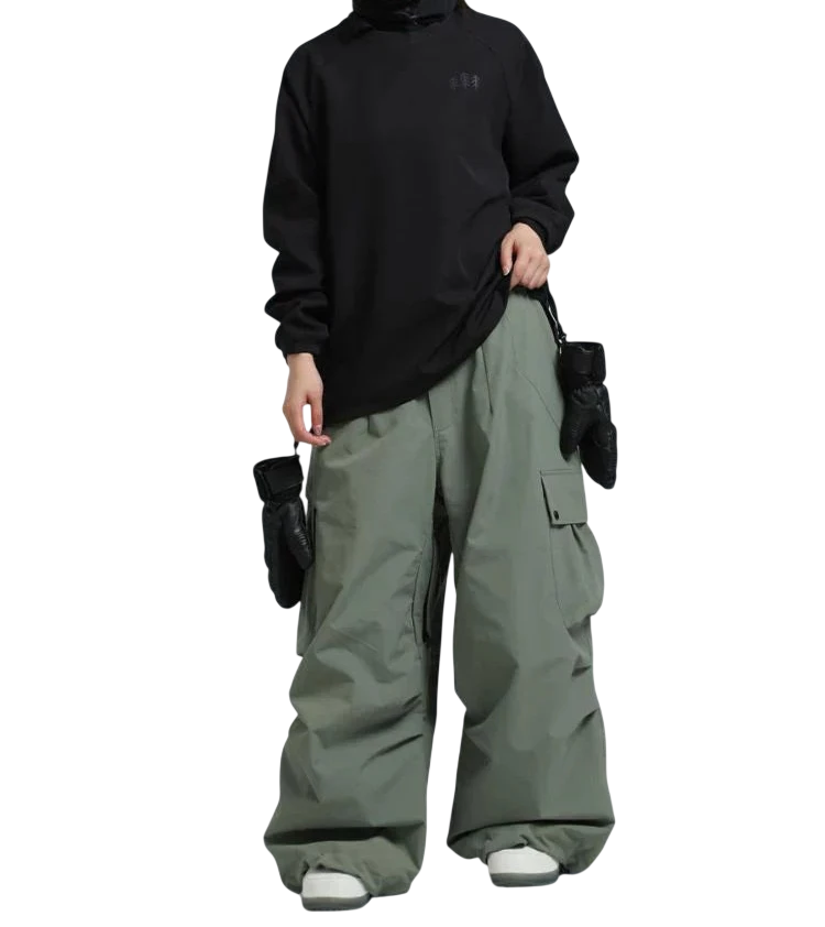 Osoba ubrana w czarną bluzę z kapturem, Arkadya Outlet spodnie zimowe męskie szeroki krój regularny na warunki zimowe w kolorze zielonym, białe buty, czarne rękawiczki i odblaskowe gogle narciarskie stoi na jednolitym szarym tle.