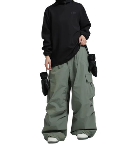 Osoba ubrana w czarną bluzę z kapturem, Arkadya Outlet spodnie zimowe męskie szeroki krój regularny na warunki zimowe w kolorze zielonym, białe buty, czarne rękawiczki i odblaskowe gogle narciarskie stoi na jednolitym szarym tle.