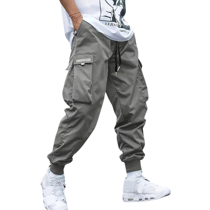 Osoba ubrana w Arkadya Outlet Męskie spodnie cargo | Dopasowana nogawka z wieloma kieszeniami, białe trampki i biały t-shirt z grafiką stoi na jednolitym jasnoszarym tle.