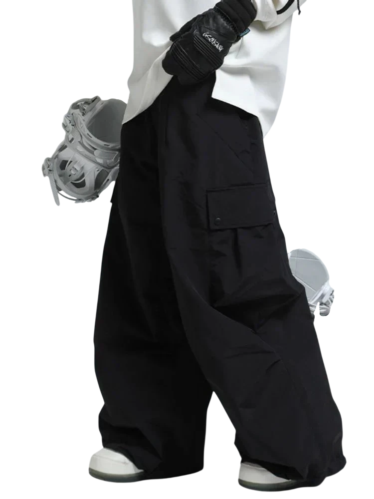 Osoba ubrana w Arkadya Outlet spodnie zimowe męskie szeroki krój regularny i białe buty śniegowe, w czarnych rękawiczkach, stoi trzymając deskę snowboardową pod jedną ręką. Obraz koncentruje się na dolnej części ciała.