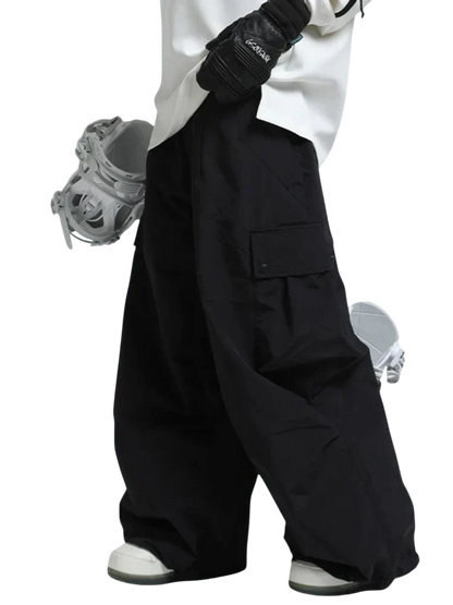 Osoba ubrana w Arkadya Outlet spodnie zimowe męskie szeroki krój regularny i białe buty śniegowe, w czarnych rękawiczkach, stoi trzymając deskę snowboardową pod jedną ręką. Obraz koncentruje się na dolnej części ciała.