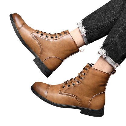 Osoba nosząca Arkadya Outlet Buty męskie wysoka cholewka z wiązaniem pełna wysokość w kolorze jasnobrązowym z czarnymi podeszwami i czarnymi dżinsami z postrzępioną rąbkiem, opierająca nogi na teksturowanym szarym tle.