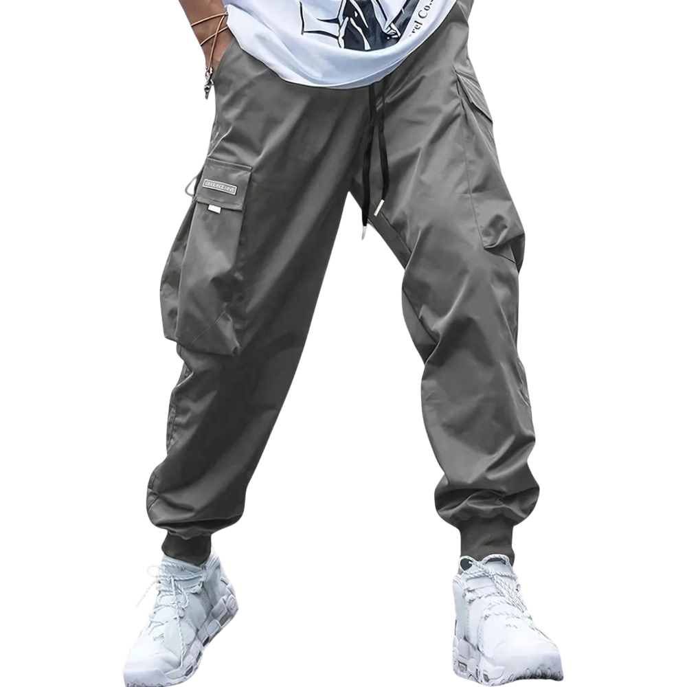 Mężczyzna ubrany w spodnie cargo Arkadya Outlet Męskie ze zwężanymi nogawkami i wieloma kieszeniami, w połączeniu z białymi trampkami i białą koszulką z grafiką, stojący na jednolitym jasnoszarym tle.