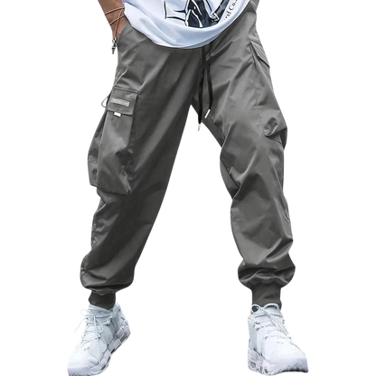 Mężczyzna ubrany w spodnie cargo Arkadya Outlet Męskie ze zwężanymi nogawkami i wieloma kieszeniami, w połączeniu z białymi trampkami i białą koszulką z grafiką, stojący na jednolitym jasnoszarym tle.