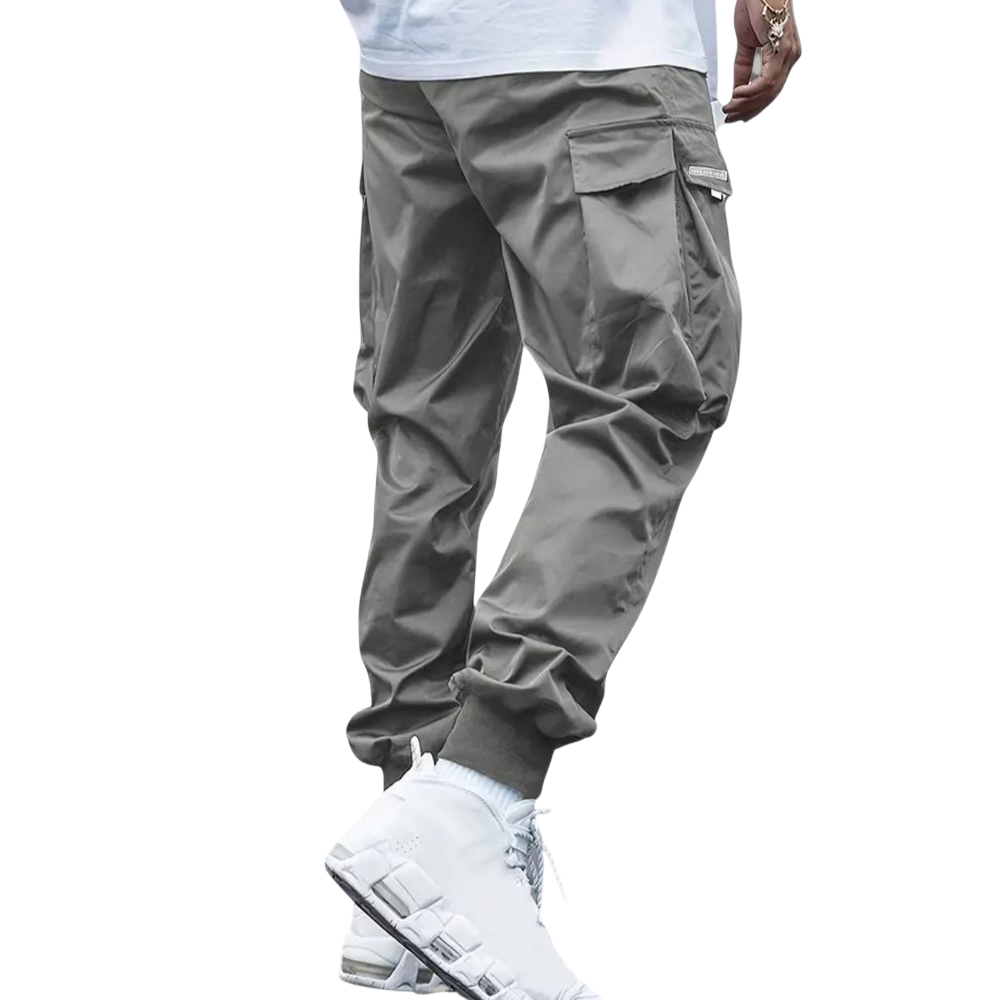 Osoba ubrana w spodnie cargo Arkadya Outlet Męskie, pokazana od pasa w dół na jasnym tle. Jasnoszare joggery mają boczne kieszenie, wąski krój i elastyczne mankiety przy kostkach.