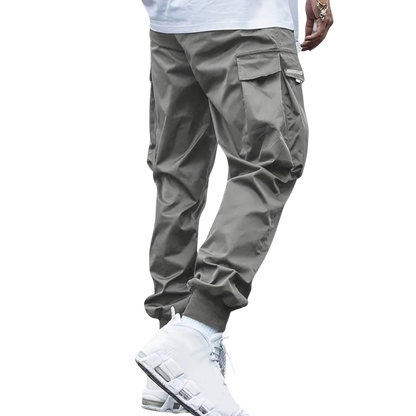 Osoba ubrana w spodnie cargo Arkadya Outlet Męskie, pokazana od pasa w dół na jasnym tle. Jasnoszare joggery mają boczne kieszenie, wąski krój i elastyczne mankiety przy kostkach.