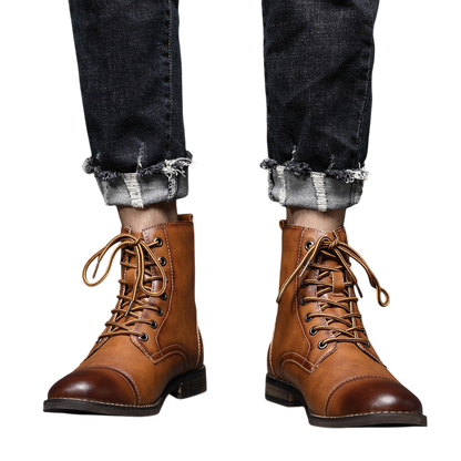 Osoba w podwiniętych ciemnych dżinsach i Arkadya Outlet Buty męskie wysoka cholewka z wiązaniem pełna wysokość stoi na wyłożonej kafelkami podłodze przy szarej ścianie.