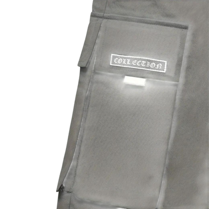 Zbliżenie na szarą kieszeń cargo z klapką i białą metką "COLLECTION" na męskim spodnie cargo marki Arkadya Outlet; posiada białą zakładkę pod metką i znajduje się na jednolitym jasnoszarym tle.
