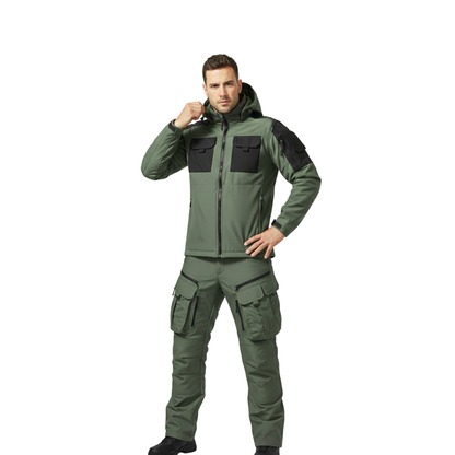 Mężczyzna pozuje pewnie na białym tle, ubrany w Arkadya Outlet zestaw odzieży outdoorowej męska kurtka i spodnie wodoodporne w kolorze oliwkowo-zielonym z czarnymi naszywkami, jedną rękę trzyma na biodrze, a drugą dotyka kaptura.