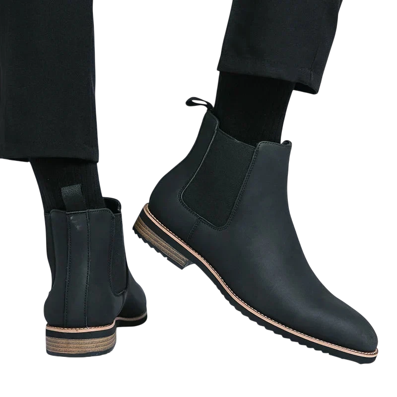 Osoba nosi Arkadya Outlet Botki Chelsea męskie zimowe styl Chelsea na chłód z czarnymi skarpetami i spodniami. Jedna stopa jest płaska, a druga uniesiona, aby pokazać bok buta i drewnianą podeszwę na białym tle.