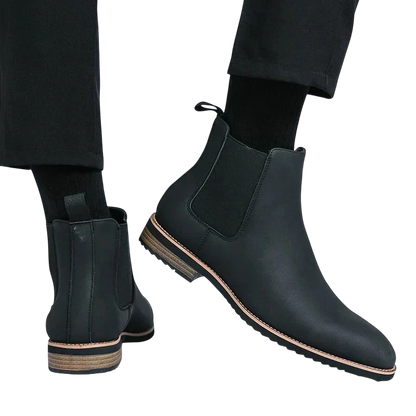 Osoba nosi Arkadya Outlet Botki Chelsea męskie zimowe styl Chelsea na chłód z czarnymi skarpetami i spodniami. Jedna stopa jest płaska, a druga uniesiona, aby pokazać bok buta i drewnianą podeszwę na białym tle.