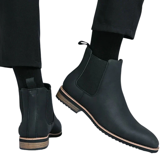 Osoba nosi Arkadya Outlet Botki Chelsea męskie zimowe styl Chelsea na chłód z czarnymi skarpetami i spodniami. Jedna stopa jest płaska, a druga uniesiona, aby pokazać bok buta i drewnianą podeszwę na białym tle.