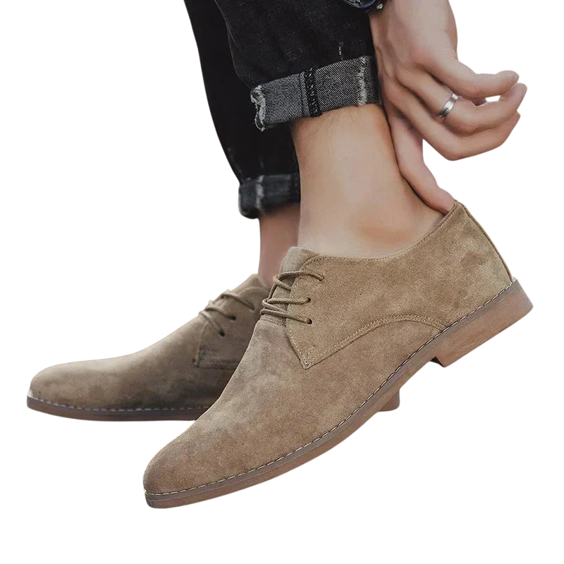 Osoba w podwiniętych czarnych dżinsach zakłada Arkadya Outlet Buty Biznesowe Męskie Płaski Obcas Zamknięty Nosek, jasnobrązowe zamszowe sznurowane buty z podeszwami z przeszyciami i klasycznym wzorem. Tło jest gładkie, białe.