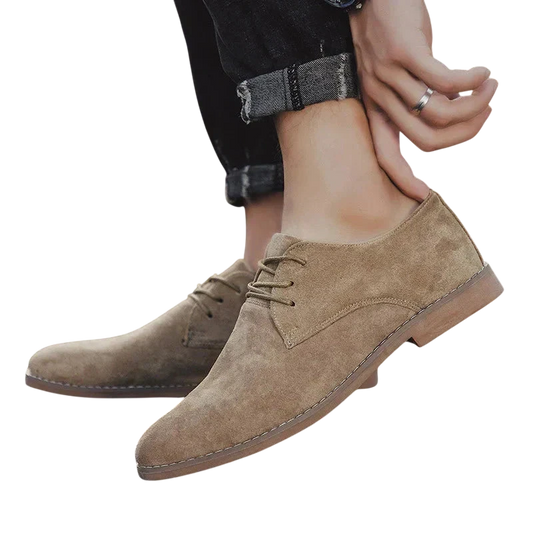 Osoba w podwiniętych czarnych dżinsach zakłada Arkadya Outlet Buty Biznesowe Męskie Płaski Obcas Zamknięty Nosek, jasnobrązowe zamszowe sznurowane buty z podeszwami z przeszyciami i klasycznym wzorem. Tło jest gładkie, białe.