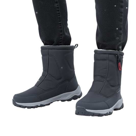 Osoba w ciemnych spodniach nosi Arkadya Outlet Męskie buty zimowe średnia wysokość wodoodporna izolacja, stojąc na drewnianej powierzchni na zewnątrz w śnieżnym krajobrazie z padającym śniegiem i drzewami w tle.