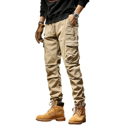 Mężczyzna w Arkadia Outlet Spodnie cargo męskie taktyczne wielokieszeniowa konstrukcja, brązowe buty robocze, czarna koszula z długim rękawem, zegarek i brązowe rękawiczki stoi na prostym białym tle.