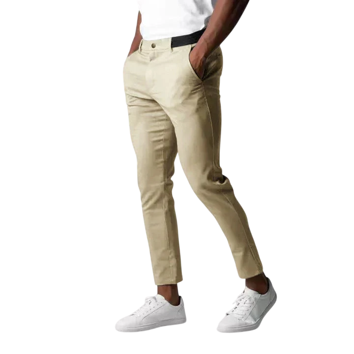 Mężczyzna stoi z rękami w kieszeniach na jasnoszarym tle, ubrany w Arkadya Outlet Spodnie chinosy męskie modny krój pełna długość użytkowy pas w kolorze beżowym, białą koszulkę i białe trampki.