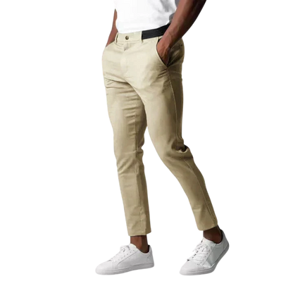 Mężczyzna stoi z rękami w kieszeniach na jasnoszarym tle, ubrany w Arkadya Outlet Spodnie chinosy męskie modny krój pełna długość użytkowy pas w kolorze beżowym, białą koszulkę i białe trampki.