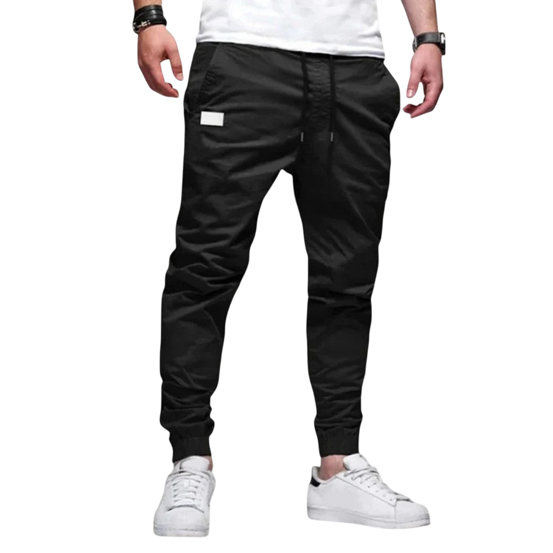 Mężczyzna modelujący Arkadya Outlet Spodnie joggerowe męskie krój slim ze ściągaczem w pasie i mankietach, biały t-shirt, białe sneakersy i czarne opaski na nadgarstki stoi na jednolitym jasnym tle. Zdjęcie przycięte poniżej ramion.