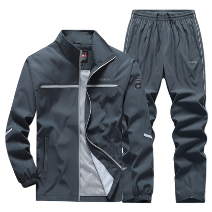 Arkadya Outlet Dres komplet męski dwuczęściowy obejmuje ciemnoszarą, zapinaną na zamek bluzę ze stójką i odblaskowymi detalami oraz elastyczne spodnie z bocznymi kieszeniami i mankietami.