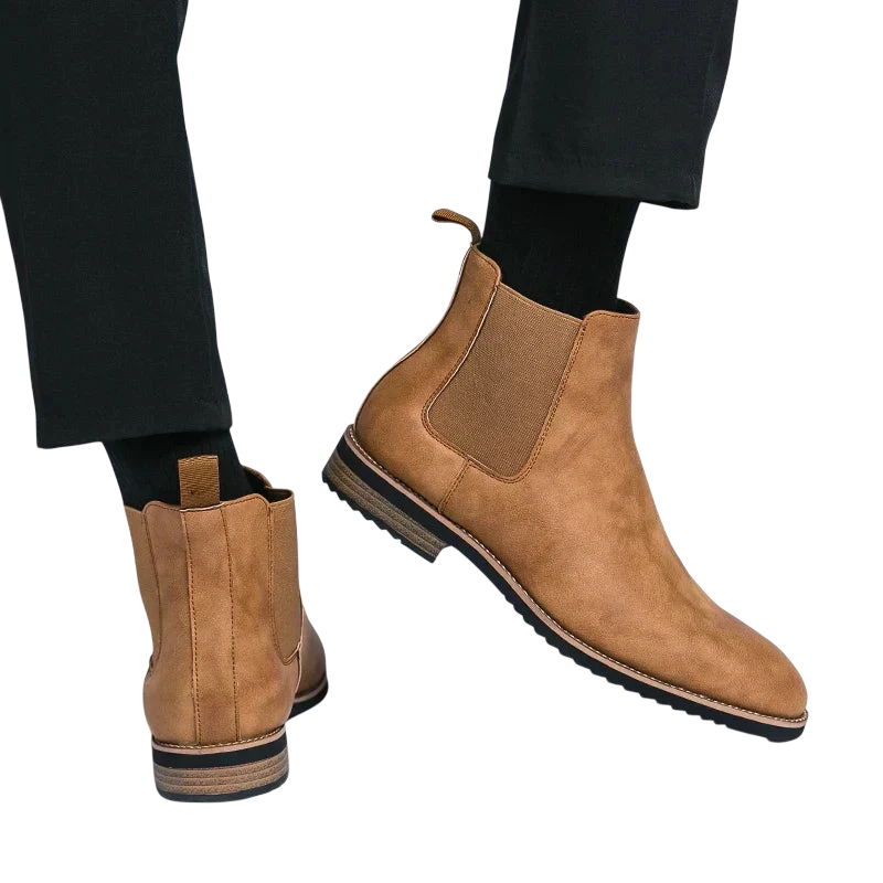 Mężczyzna ubrany w Arkadya Outlet Botki Chelsea męskie zimowe styl Chelsea na chłód z brązowego zamszu z ciemnymi podeszwami i czarnymi elastycznymi panelami bocznymi, w połączeniu z czarnymi skarpetami i spodniami, stoi na białym tle.