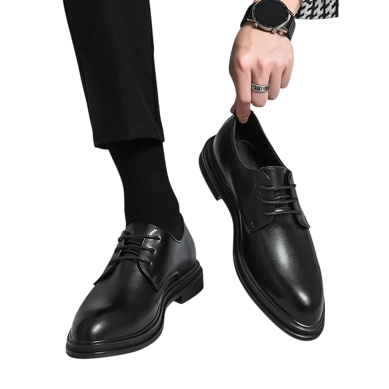 Osoba w czarnych spodniach i skarpetkach trzyma parę Arkadya Outlet Buty Wizytowe Męskie Sznurowane Oxfordy Spiczasty Nosek, z jednym błyszczącym czarnym butem na podłodze, a drugim uniesionym w dłoni.