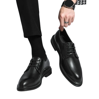 Osoba w czarnych spodniach i skarpetkach trzyma parę Arkadya Outlet Buty Wizytowe Męskie Sznurowane Oxfordy Spiczasty Nosek, z jednym błyszczącym czarnym butem na podłodze, a drugim uniesionym w dłoni.