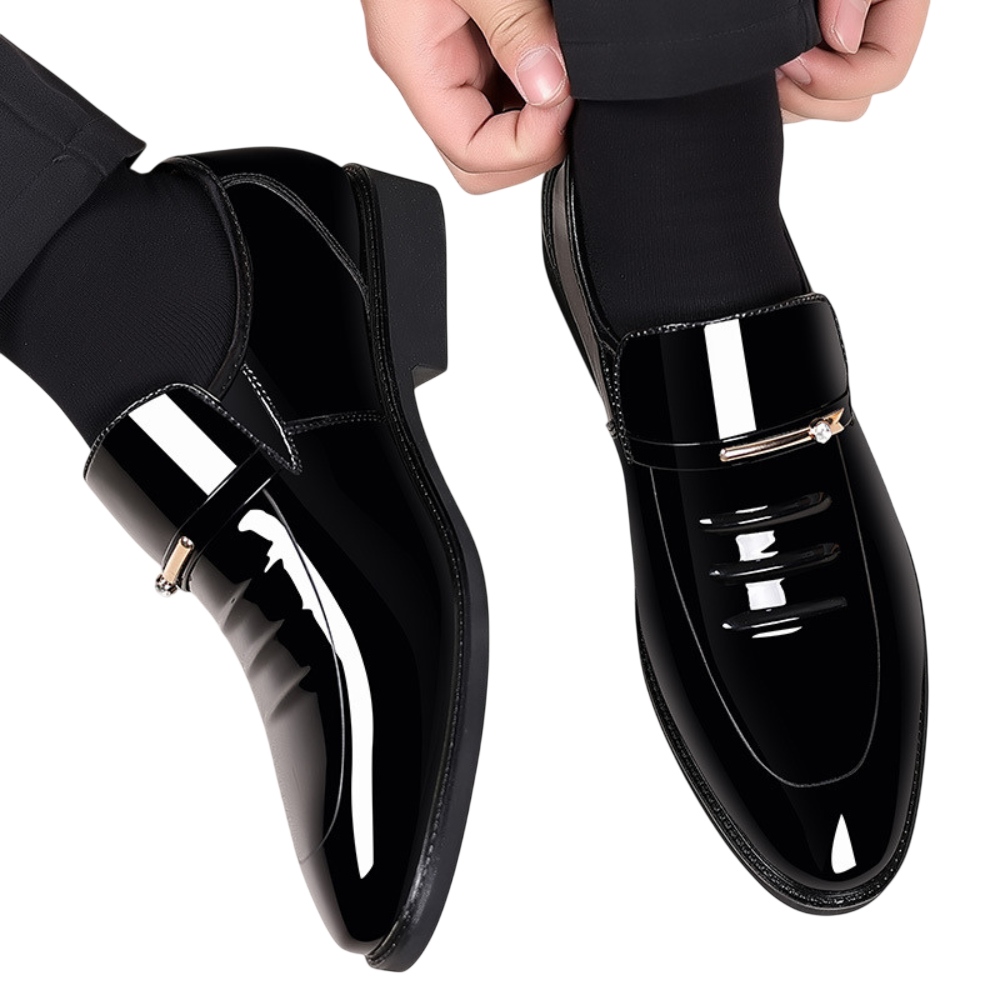 Osoba nosząca Arkadya Outlet Mokasyny Wizytowe Męskie Wsuwany Szpiczasty Nosek w błyszczącej czerni z czarnymi skarpetkami, regulująca mankiet spodni. Błyszczące buty posiadają ozdobny metalowy akcent na cholewce.