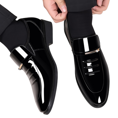 Osoba nosząca Arkadya Outlet Mokasyny Wizytowe Męskie Wsuwany Szpiczasty Nosek w błyszczącej czerni z czarnymi skarpetkami, regulująca mankiet spodni. Błyszczące buty posiadają ozdobny metalowy akcent na cholewce.