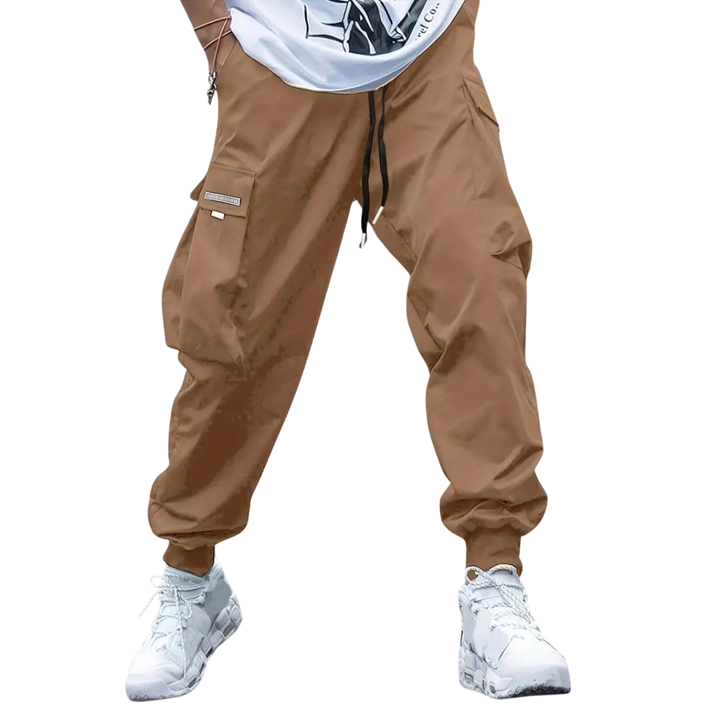 Osoba ubrana w spodnie cargo z Arkadia Outlet Męskie ze zwężanymi nogawkami i wieloma kieszeniami, w połączeniu z częściowo widoczną białą koszulką z grafiką i białymi trampkami, stoi na jednolitym jasnoszarym tle.