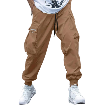 Osoba ubrana w spodnie cargo z Arkadia Outlet Męskie ze zwężanymi nogawkami i wieloma kieszeniami, w połączeniu z częściowo widoczną białą koszulką z grafiką i białymi trampkami, stoi na jednolitym jasnoszarym tle.