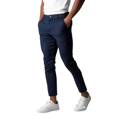 Osoba nosząca Arkadya Outlet Spodnie chinosy męskie o modnym kroju i pełnej długości, białą koszulkę i białe sneakersy stoi z rękami w kieszeniach na jasnoszarym tle. Głowa osoby nie jest widoczna na zdjęciu.