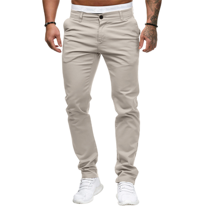 Na szarym tle stoi mężczyzna ubrany w Arkadya Outlet Męskie spodnie w dopasowanym kroju z prostą nogawką, białą koszulę, białe sneakersy i czarne bransoletki. Twarz nie jest widoczna.