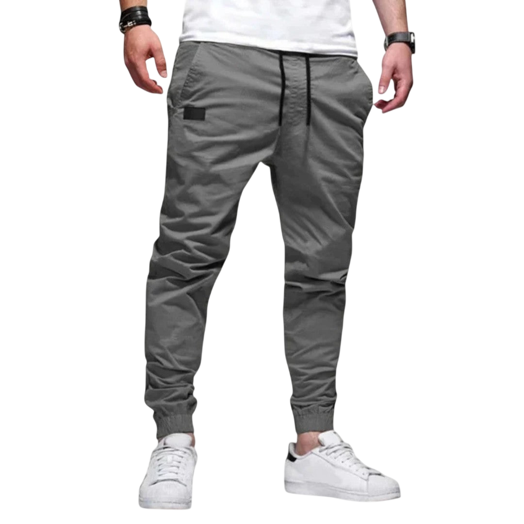 Mężczyzna nosi Arkadya Outlet Spodnie joggerowe męskie krój slim ze ściągaczem w pasie i mankietach, w stylizacji z białym t-shirtem, białymi sneakersami, czarną bransoletką i zegarkiem na rękę, na jednolitym jasnoszarym tle.