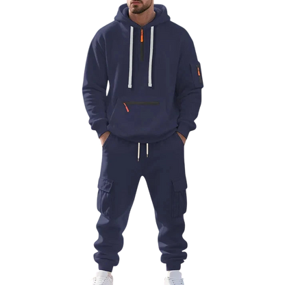 Osoba w granatowym Arkadya Outlet Męski dres wyściełane wnętrze dwuczęściowy komplet z zamkiem, z pasującymi cargo spodnie dresowe, stoi przy kamiennej fontannie w parku w słoneczny dzień. Twarz nie jest widoczna.