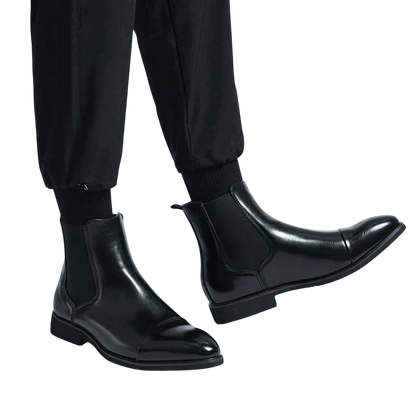 Osoba nosząca Arkadya Outlet Botki Chelsea Męskie Antypoślizgowa Zamknięta Konstrukcja w błyszczącej czerni z czarnymi spodniami, stojąca na prostym białym tle.