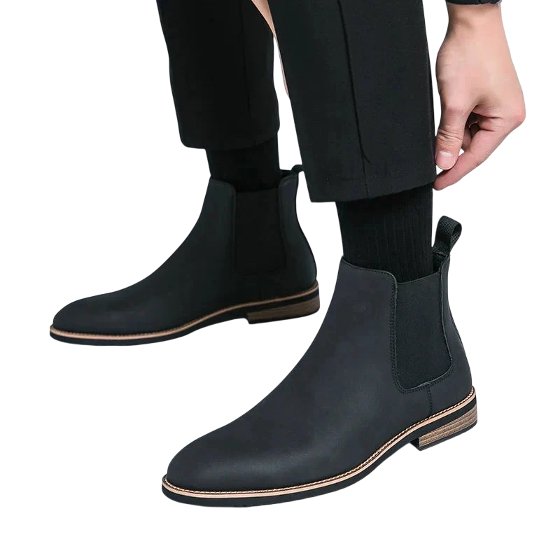 Mężczyzna w Arkadia Outlet Botki Chelsea męskie zimowe styl Chelsea na chłód z elastycznymi panelami bocznymi, brązowymi podeszwami i zakładką z tyłu, dopasowuje czarną nogawkę spodni do czarnych skarpet. Tło jest białe.