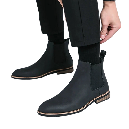 Mężczyzna w Arkadia Outlet Botki Chelsea męskie zimowe styl Chelsea na chłód z elastycznymi panelami bocznymi, brązowymi podeszwami i zakładką z tyłu, dopasowuje czarną nogawkę spodni do czarnych skarpet. Tło jest białe.