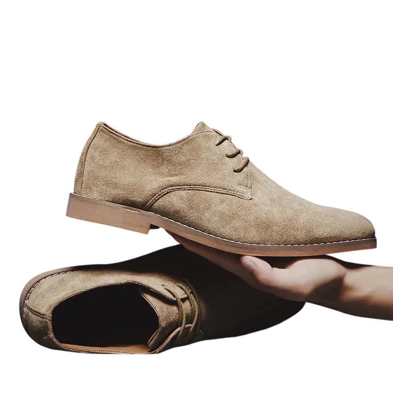 Ręka trzyma jasnobrązowe zamszowe oxfordy z Arkadya Outlet-Buty Biznesowe Męskie Płaski Obcas Zamknięty Nosek, podczas gdy pasująca para spoczywa płasko na białym tle.
