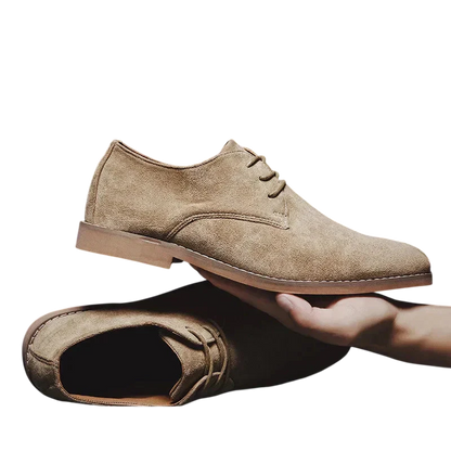 Ręka trzyma jasnobrązowe zamszowe oxfordy z Arkadya Outlet-Buty Biznesowe Męskie Płaski Obcas Zamknięty Nosek, podczas gdy pasująca para spoczywa płasko na białym tle.