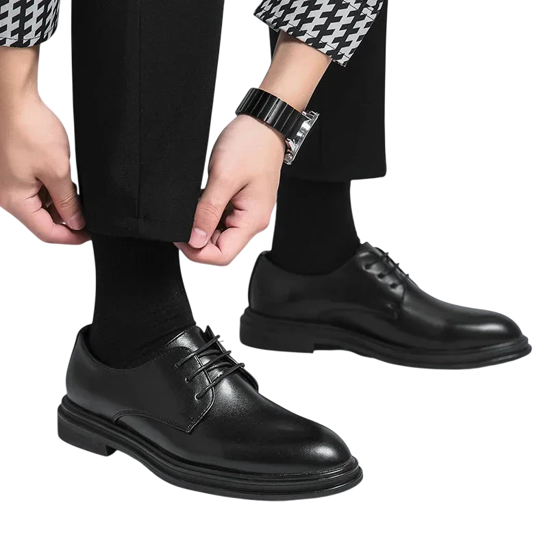 Osoba w czarnych spodniach, czarnych skarpetkach i koszuli w pepitkę reguluje mankiet, nosząc Buty Wizytowe Męskie Sznurowane Oxfordy Spiczasty Nosek z Outletu Arkadya na zwykłym białym tle.