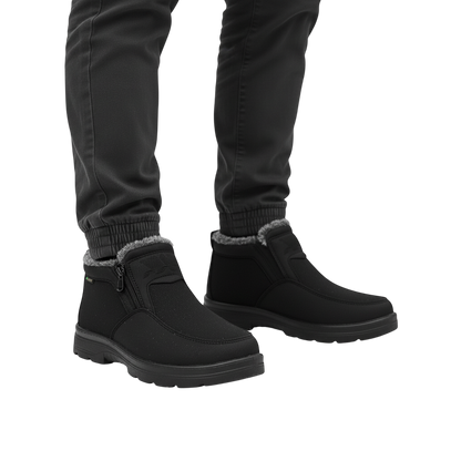Osoba w ciemnych spodniach stoi na jasnoszarej powierzchni, ubrana w Arkadya Outlet Męskie buty wodoodporne antypoślizgowe zimowe obuwie z podszewką ze sztucznego futra i bocznymi zamkami. Czarne buty mają antypoślizgową podeszwę i wodoodporną konstrukcję.