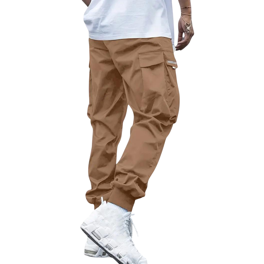 Model pokazuje tylko dolną połowę, ubrany w Męskie spodnie cargo marki Arkadya Outlet - brązowe spodnie jogger z dopasowaną nogawką i wieloma kieszeniami - w połączeniu z białym t-shirtem i białymi trampkami na jasnoszarym tle.