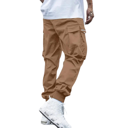 Model pokazuje tylko dolną połowę, ubrany w Męskie spodnie cargo marki Arkadya Outlet - brązowe spodnie jogger z dopasowaną nogawką i wieloma kieszeniami - w połączeniu z białym t-shirtem i białymi trampkami na jasnoszarym tle.