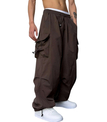 Osoba modeluje Męskie Spodnie cargo Arkadya Outlet - luźne, ciemnobrązowe spodnie z szerokimi nogawkami i wieloma kieszeniami - w połączeniu z białymi trampkami. Ich tors jest nagi, ukazując widoczny tatuaż na ramieniu, osadzony na prostym tle.