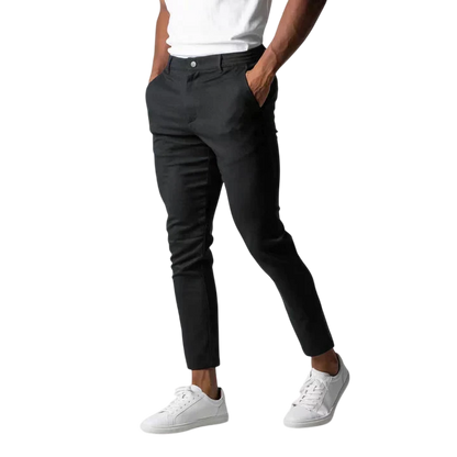 Osoba w białym t-shircie, Arkadya Outlet spodnie chinos męskie modny krój pełna długość użytkowy pas w czarnym slim-fit i białe trampki stoi z rękami w kieszeniach na jednolitym szarym tle; górna część ciała jest przycięta.