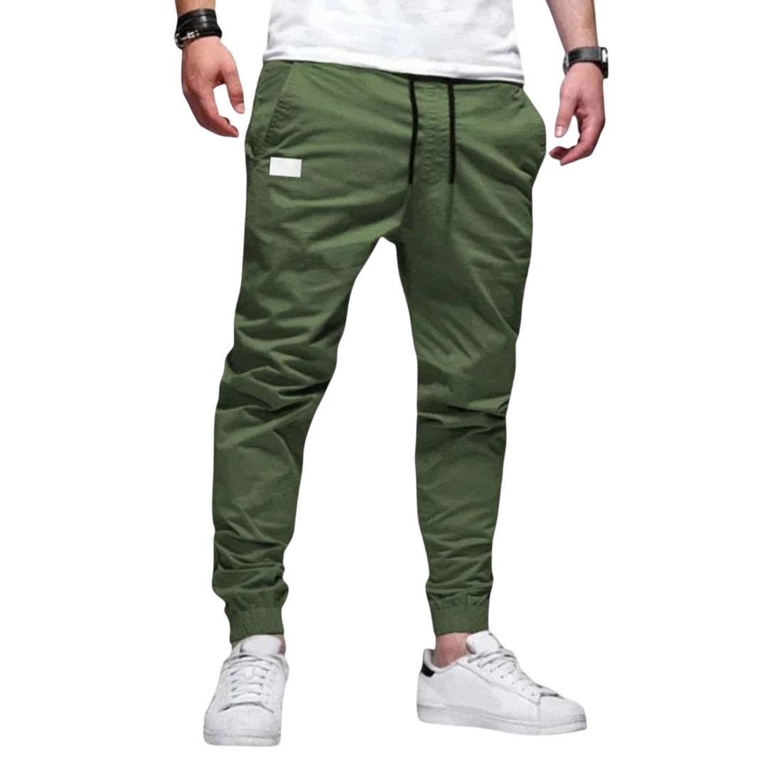 Osoba ma na sobie spodnie męskie joggerowe Arkadya Outlet Spodnie slim fit ze ściągaczem w pasie i mankietach, białymi naszywkami, białymi sneakersami, białym t-shirtem i czarnymi akcesoriami na nadgarstek na jednolitym jasnoszarym tle.