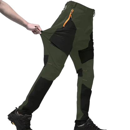 Osoba rozciąga bok swoich zielono-czarnych Spodnie Terenowe Męskie Wodoodporne Wiatroszczelne Regulowane z Outletu Arkadya, podkreślając elastyczny materiał - idealne spodnie outdoorowe, noszone z butami turystycznymi na białym tle.